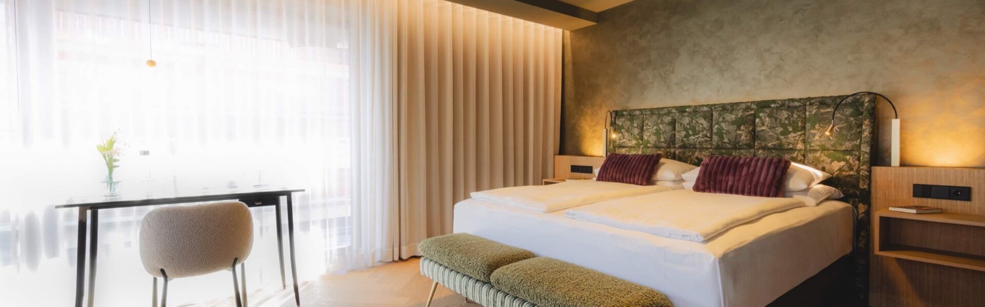 Hotel Schladming Design Suite im nōa Schladming mit Eichen-Fischgrätboden, Kunst-Headboard, Schreibtisch und großem Fenster.