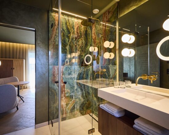 Luxuriöses Badezimmer im nōa Hotel Schladming mit Naturstein, goldenen Armaturen und moderner Regendusche